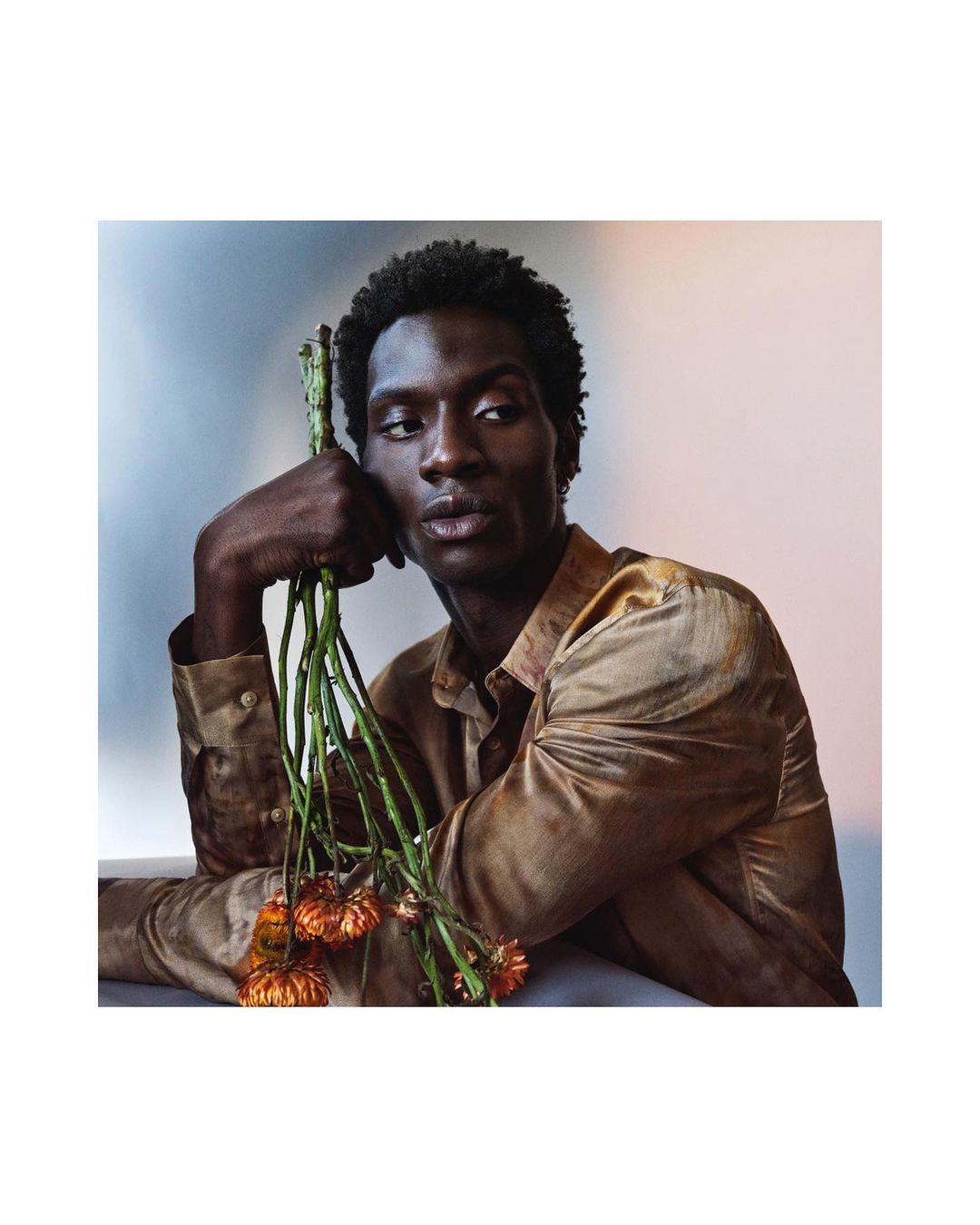 Portrait of a Black male model in a bronze shirt holding a bouquet of flowers against a gradient light blue to pale peach background.