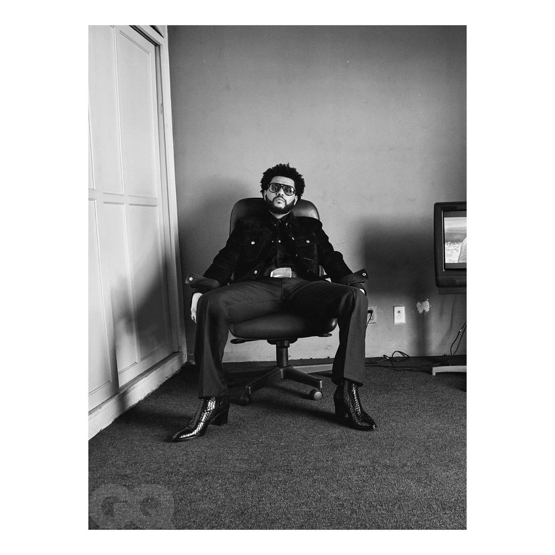 The Weeknd is sitting on a black chair. He is wearing a black jacket, dark pants, and black shoes. He has sunglasses on.