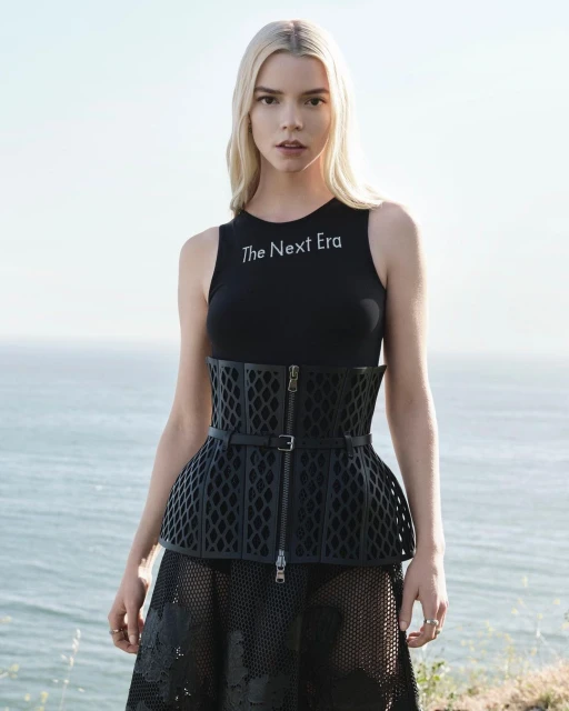 Anya Taylor-Joy poses in a black top, corset, and skirt with the ocean in the background, showcasing fashion and celebrity style.