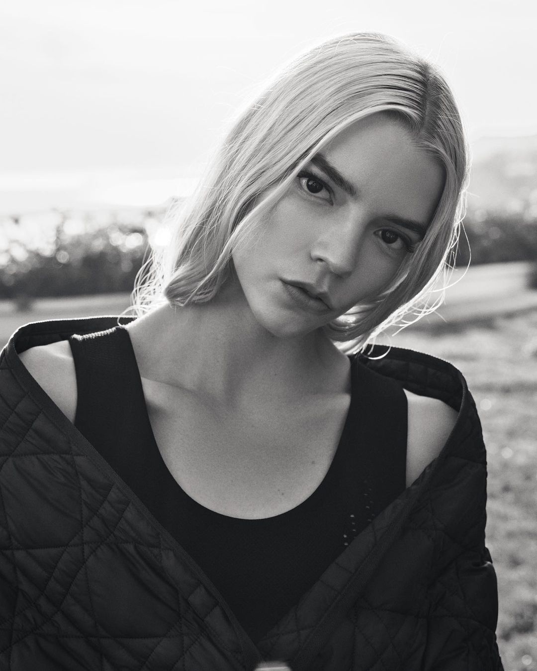 Black and white portrait of a woman with fair hair, wearing a dark top and quilted jacket, posing outdoors in natural light.