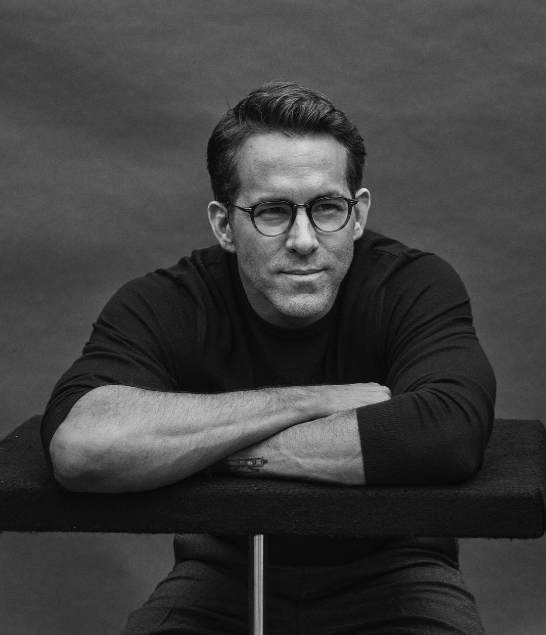Monochrome portrait of Ryan Reynolds wearing round eyeglasses and a dark long-sleeve shirt, leaning arms on a ledge.