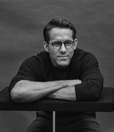 Monochrome portrait of Ryan Reynolds wearing round eyeglasses and a dark long-sleeve shirt, leaning arms on a ledge.