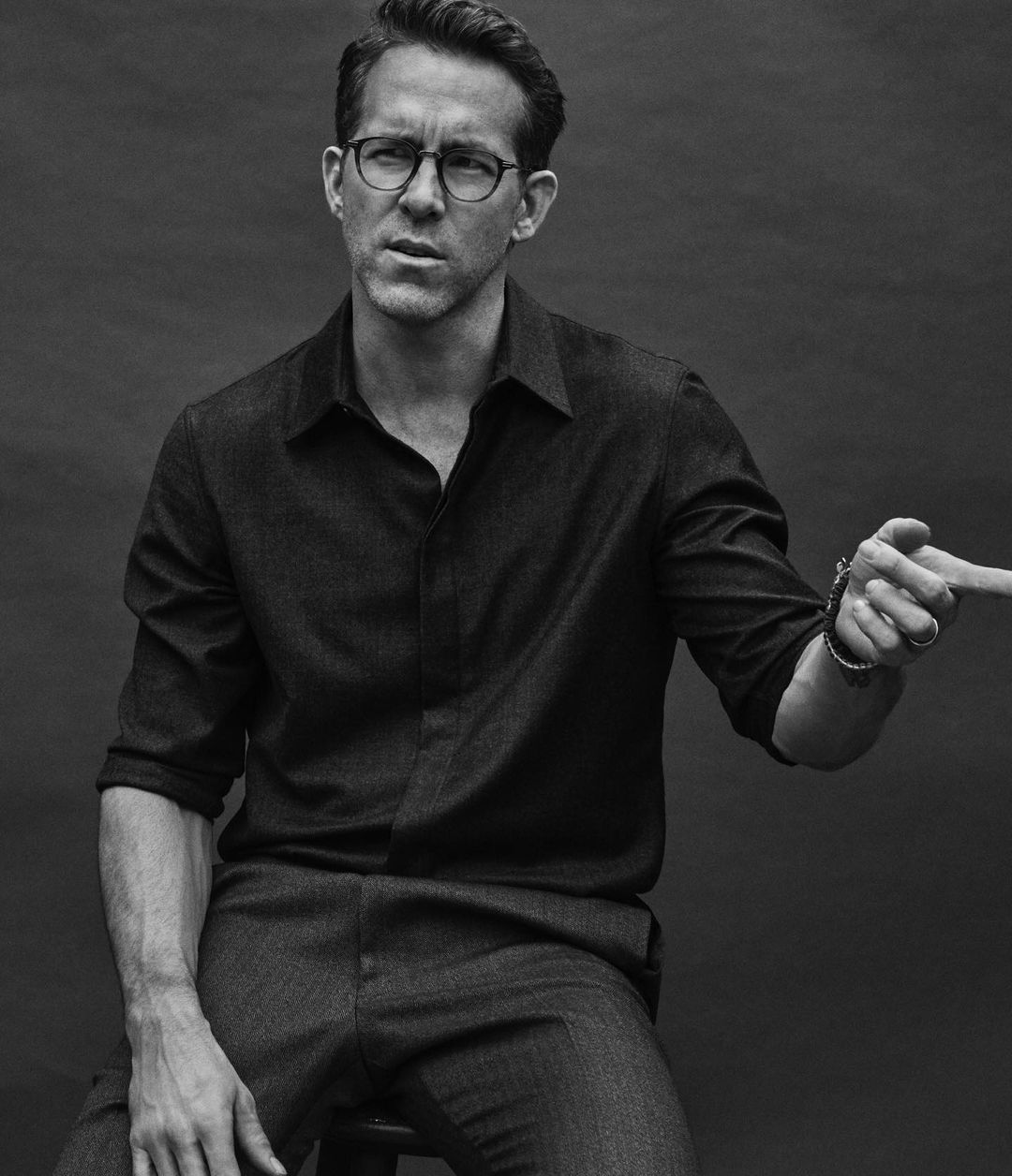 Ryan Reynolds poses in a black-and-white medium shot, seated indoors, wearing eyeglasses and pointing to the right with a serious expression.