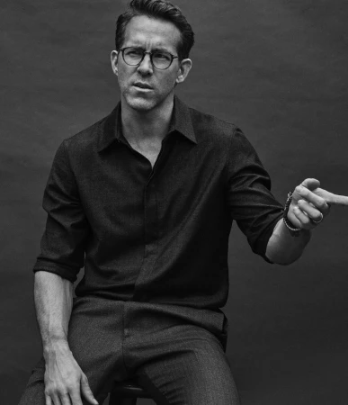 Ryan Reynolds poses in a black-and-white medium shot, seated indoors, wearing eyeglasses and pointing to the right with a serious expression.