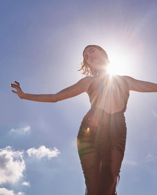 Adult female model in a dark sheer dress poses with arms outstretched against a sky with bright sun glare; natural lighting and wide shot.