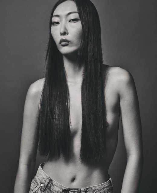 Portrait of an adult Asian woman in light denim jeans, with long dark hair against a dark grey background in a studio setting.
