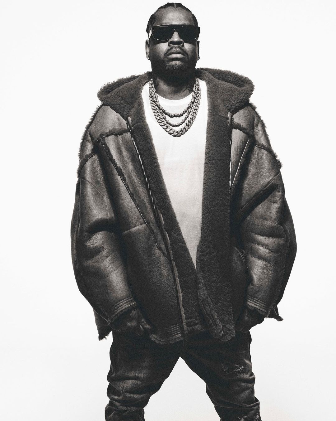 Monochrome portrait of an adult male wearing sunglasses, a shearling hooded coat, a white t-shirt, and a chunky chain necklace. 