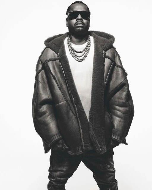 Monochrome portrait of an adult male wearing sunglasses, a shearling hooded coat, a white t-shirt, and a chunky chain necklace. 