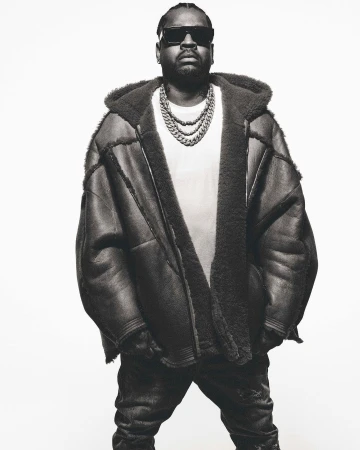 Monochrome portrait of an adult male wearing sunglasses, a shearling hooded coat, a white t-shirt, and a chunky chain necklace. 