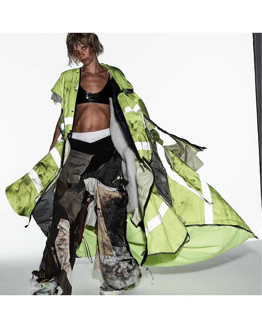Fashion photo of a woman with short blond hair wearing a black bra, layered clothing, and patchwork pants against a white background.