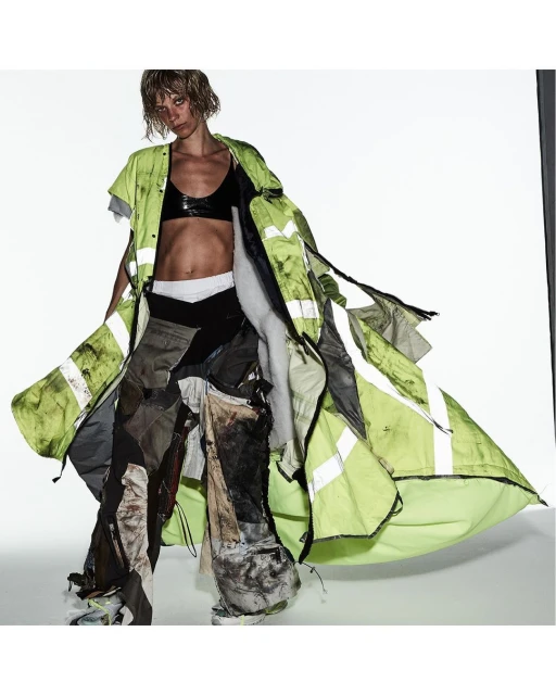 Fashion photo of a woman with short blond hair wearing a black bra, layered clothing, and patchwork pants against a white background.