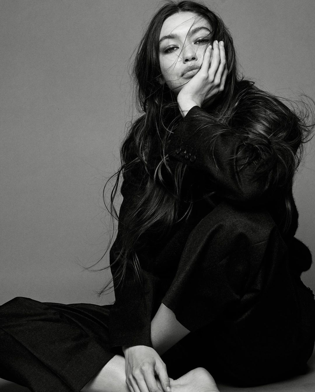 Portrait of a woman with long dark hair wearing a black blazer and pants, sitting against a grey background in black and white.