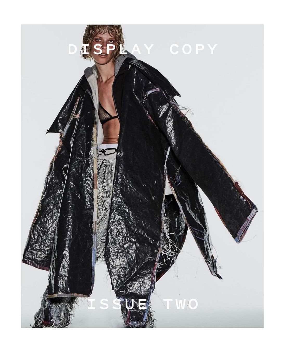 A woman models a deconstructed black vinyl trench coat over layered apparel in a full shot against a stark white backdrop.