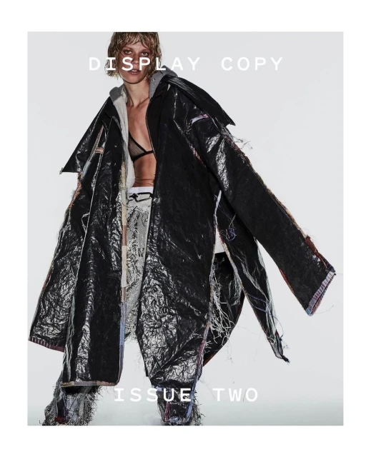 A woman models a deconstructed black vinyl trench coat over layered apparel in a full shot against a stark white backdrop.