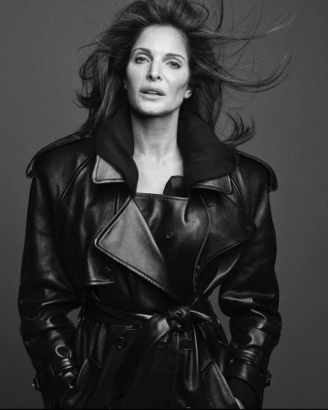 Black and white portrait of a woman with flowing hair wearing a dark leather trench coat against a plain background.