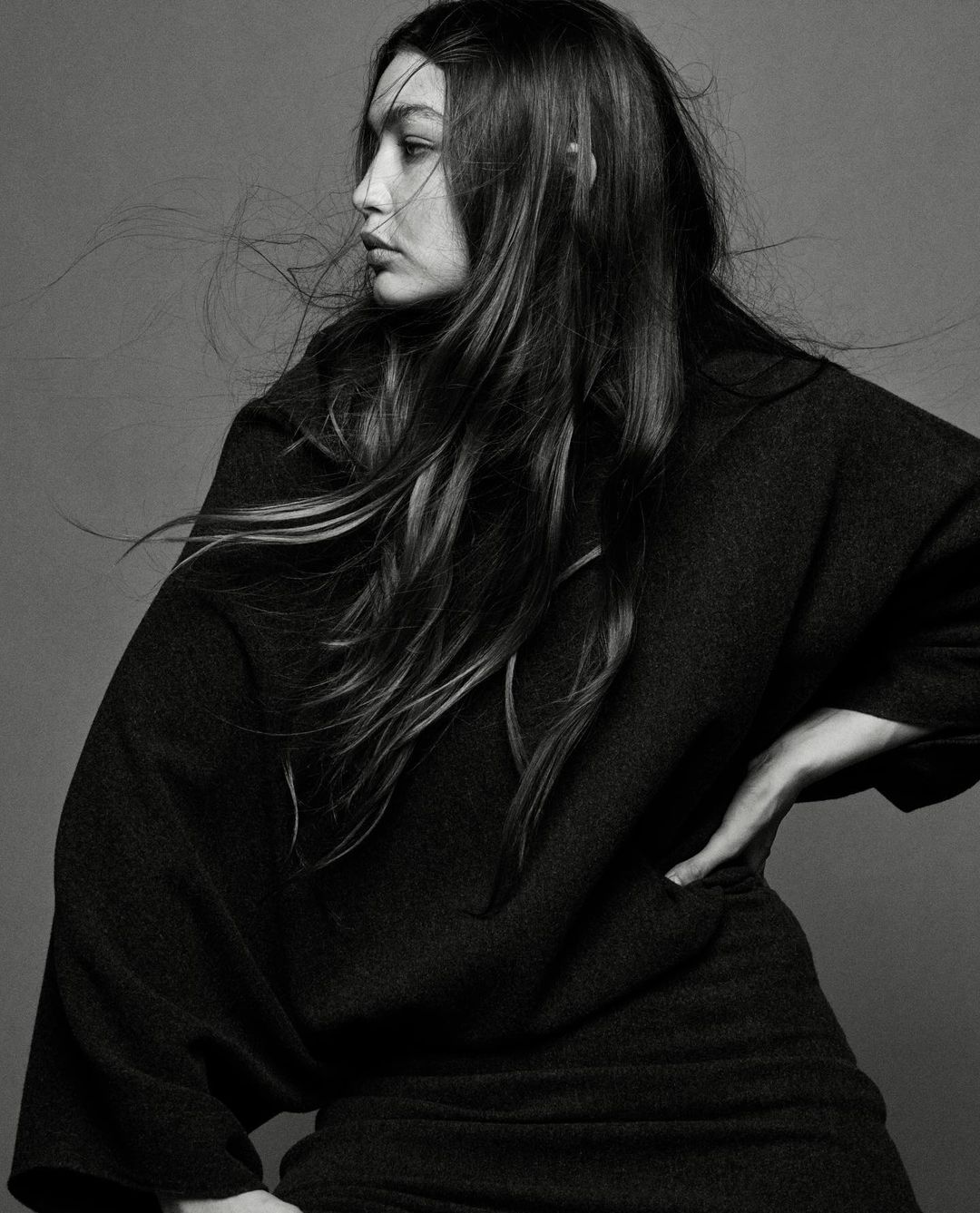 Monochrome portrait of a woman with long brown hair, wearing a dark sweater, posing against a plain grey background in a photo studio.