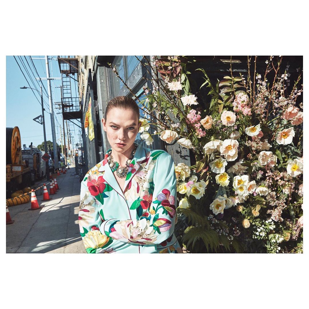 A female model poses in a floral jacket and necklace, standing on an urban street with flowers to her side, on a sunny day.