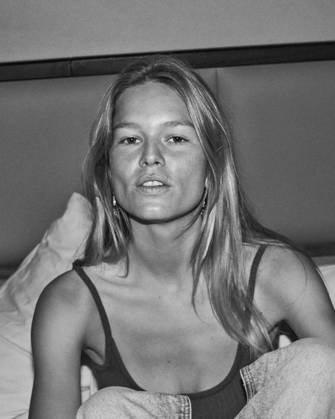Black and white portrait of a woman with long hair seated against a cushioned headboard wearing a tank top.