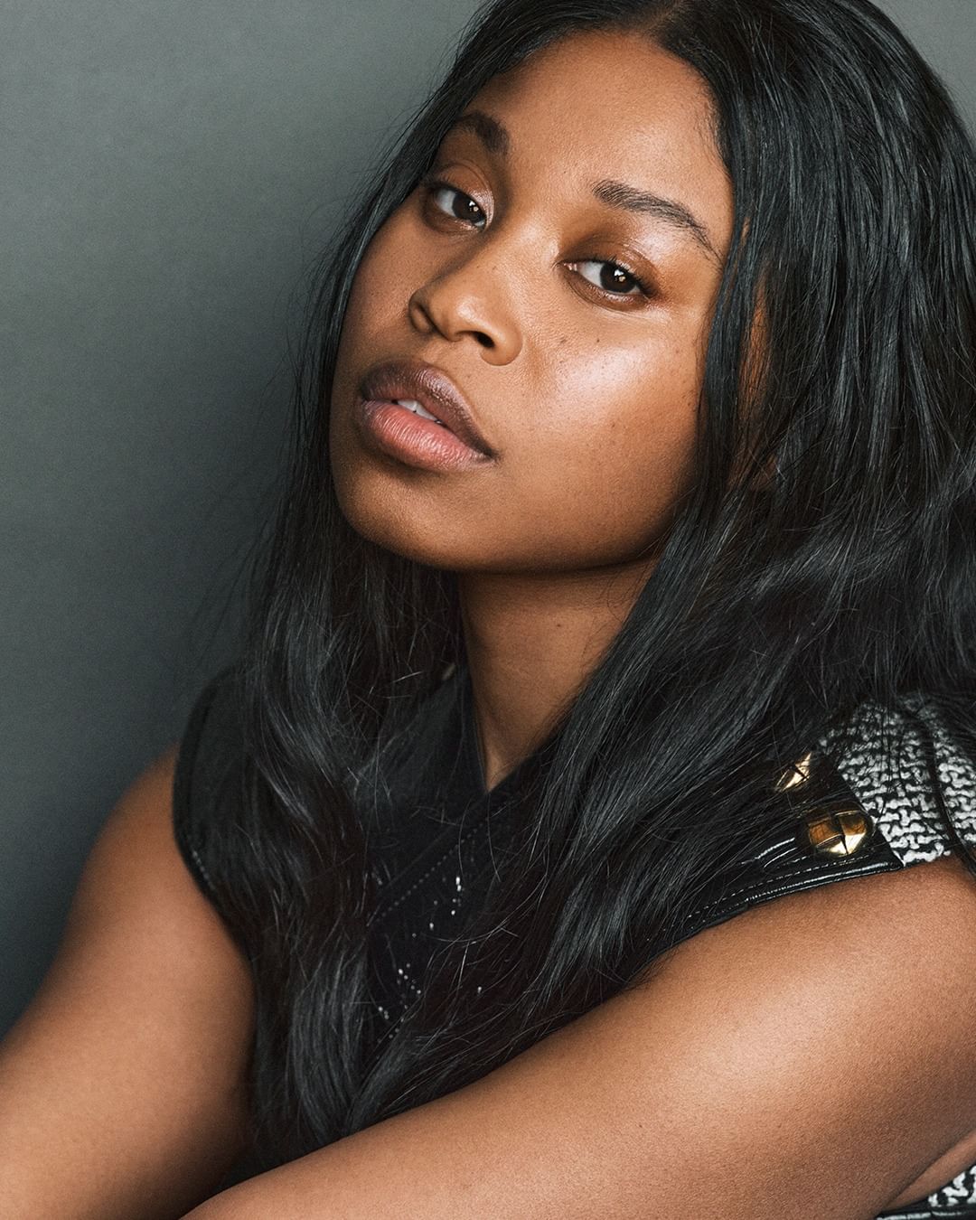 Portrait of a black woman with long dark hair wearing a sleeveless patterned garment against a dark grey background