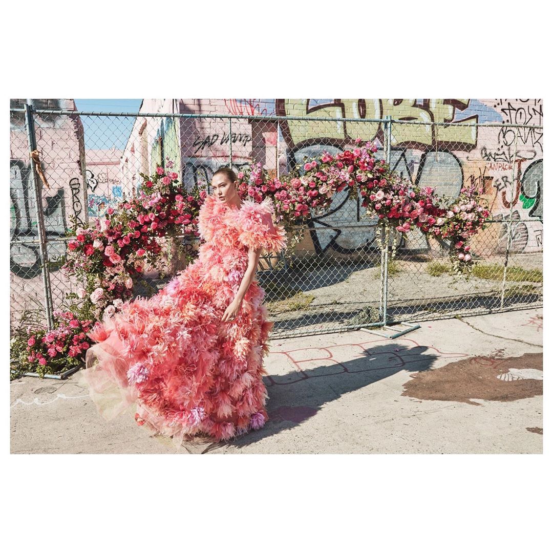 A woman in a large feathered pink gown stands in front of a graffiti-covered wall with pink flower decorations on a chain-link fence.