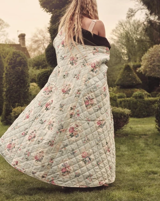 Adult woman standing outdoors in garden wearing a light blue quilted floral pattern gown and a black sweater in a tranquil setting.