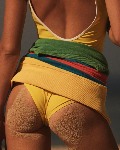 Close-up shot of a woman's back and bottom, covered in sand, wearing a yellow swimsuit and a colorful sweater wrapped around her waist.