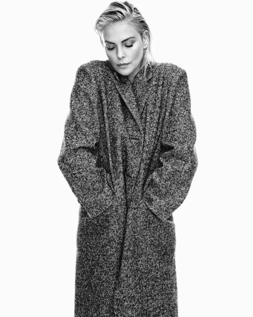 Black and white portrait of an adult female in a long, patterned coat against a white backdrop, looking down with wet, slicked-back hair.