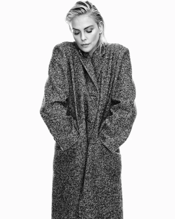 Black and white portrait of an adult female in a long, patterned coat against a white backdrop, looking down with wet, slicked-back hair.