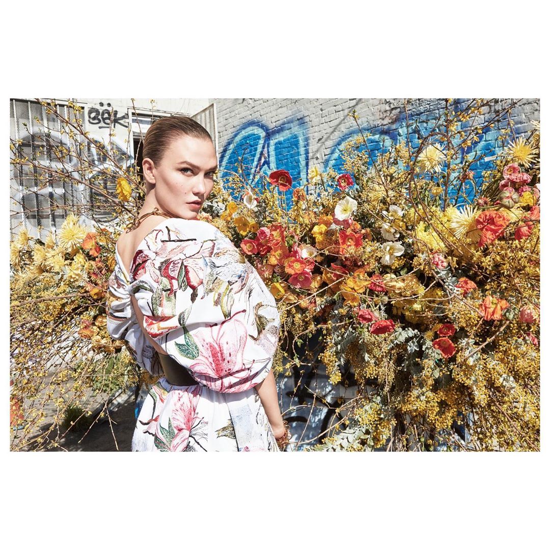 A fashion shot of a woman in a floral dress standing in front of a graffiti-covered brick wall and a large floral arrangement.