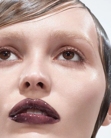 Close-up portrait of a woman with slicked-back hair and glossy plum lipstick against a bright, white background showcasing beauty.
