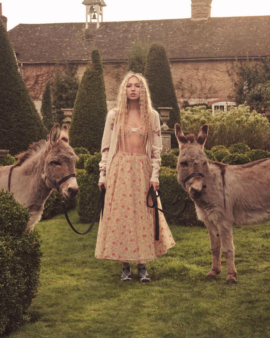 Woman stands in a garden with two donkeys, wearing a floral dress, sweater, and shoes. Building, hedges, and lawn in the background.