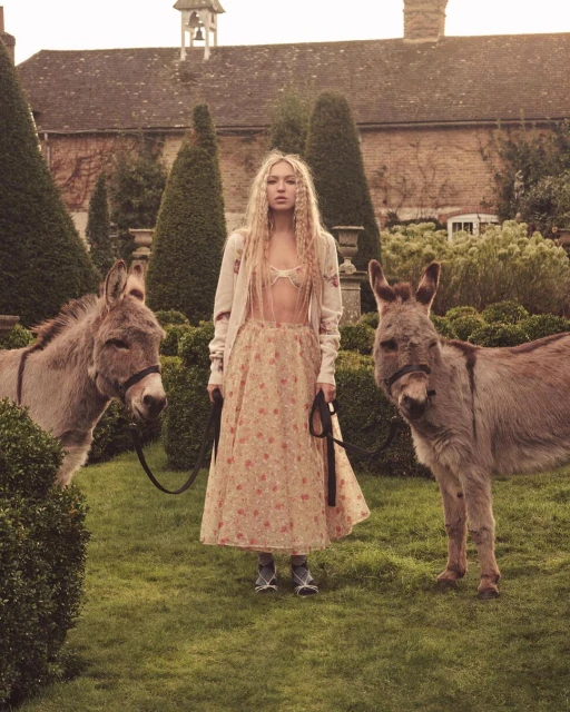 Woman stands in a garden with two donkeys, wearing a floral dress, sweater, and shoes. Building, hedges, and lawn in the background.