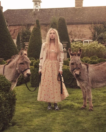 Woman stands in a garden with two donkeys, wearing a floral dress, sweater, and shoes. Building, hedges, and lawn in the background.