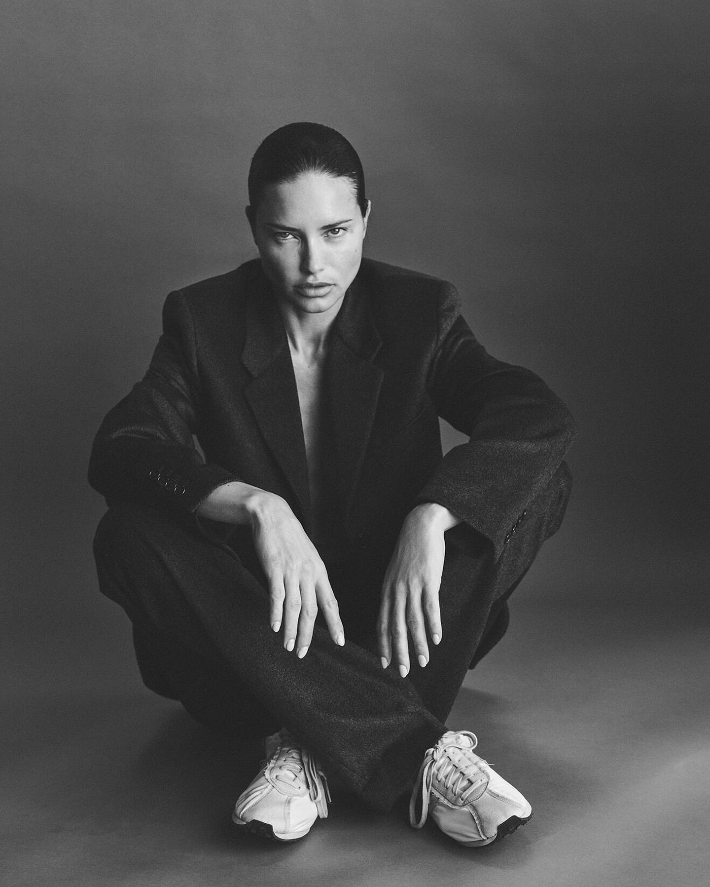 Portrait of a woman seated wearing an oversized dark suit jacket, trousers, and white sneakers against a grey backdrop