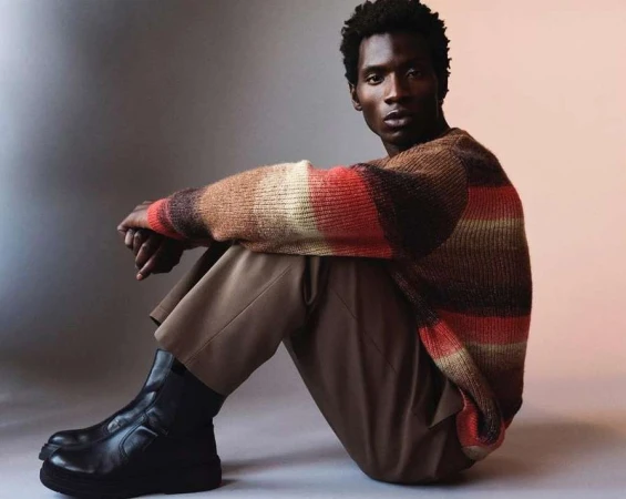 Black man wearing a striped sweater and brown trousers seated against a neutral studio backdrop