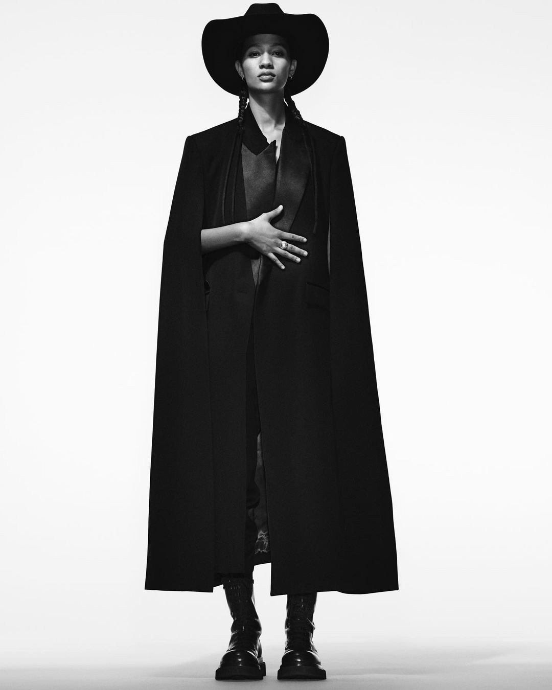 Full-length portrait of a young adult wearing a black cowboy hat, long cape, and boots against a white backdrop in a studio shot.
