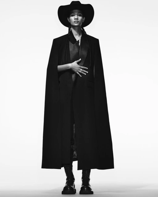 Full-length portrait of a young adult wearing a black cowboy hat, long cape, and boots against a white backdrop in a studio shot.