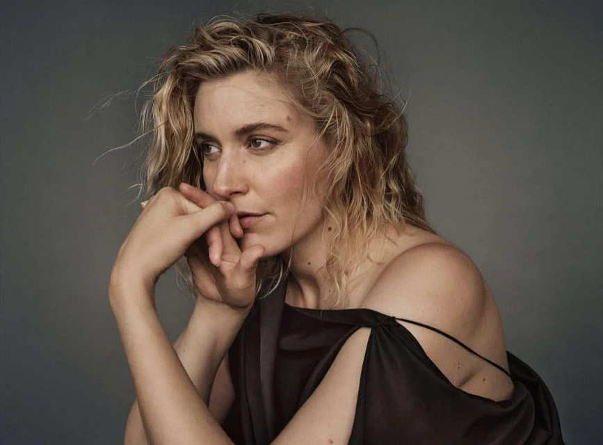 Portrait of a blonde woman wearing a black top, looking off-camera with a thoughtful expression, against a gray studio backdrop.