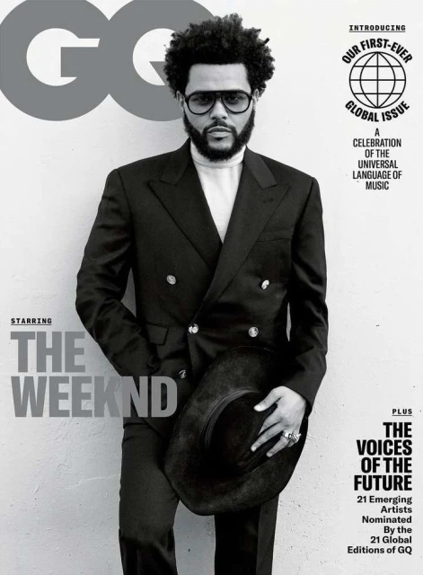 The Weeknd poses on a GQ magazine cover in a black double-breasted suit and glasses, holding a wide-brimmed black hat against a gray backdrop.