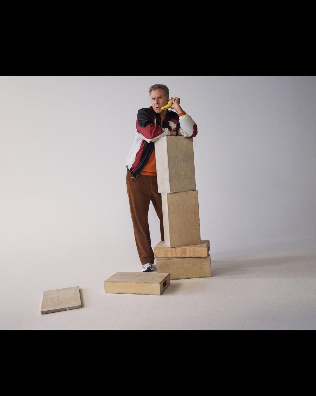 Adult male in studio, leaning on stacked concrete blocks while eating a banana, wearing a retro-style jacket and brown corduroy pants.