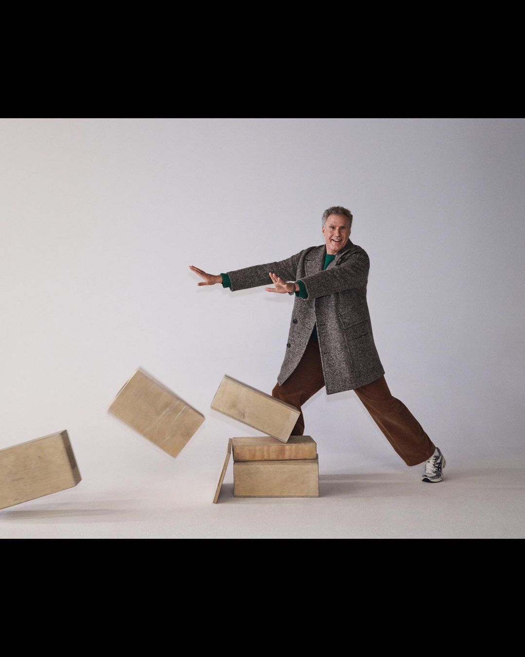 Adult male in overcoat balancing on boxes in a studio setting, wearing brown pants and sneakers with outstretched arms in studio lighting.