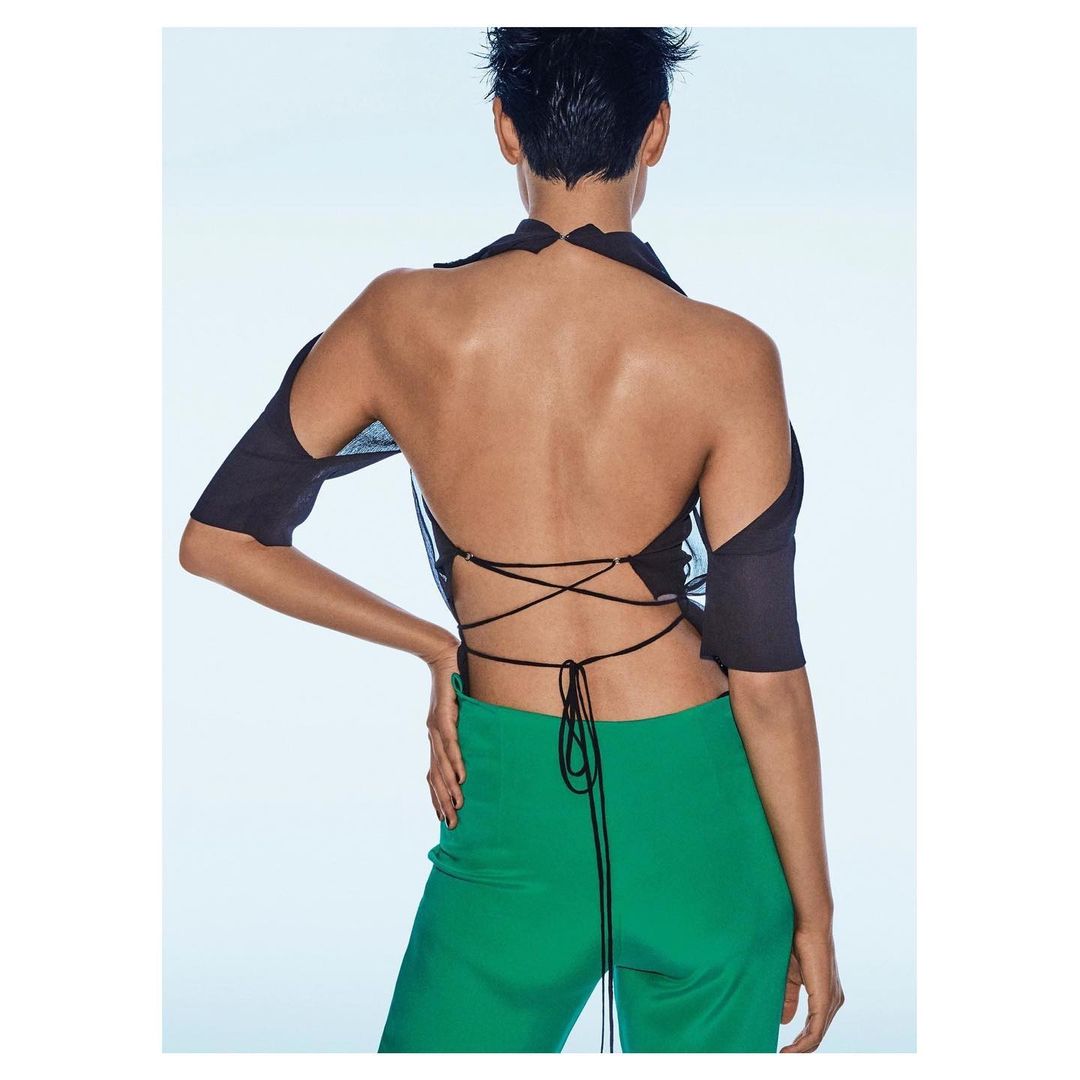 Back view of a fashion model with short hair, wearing a black top with back ties and green pants, set against a light background.