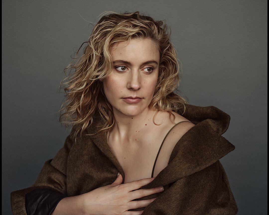 Portrait of a woman with blond curly hair wearing a brown coat, captured in a photo studio setting with a neutral gray background.