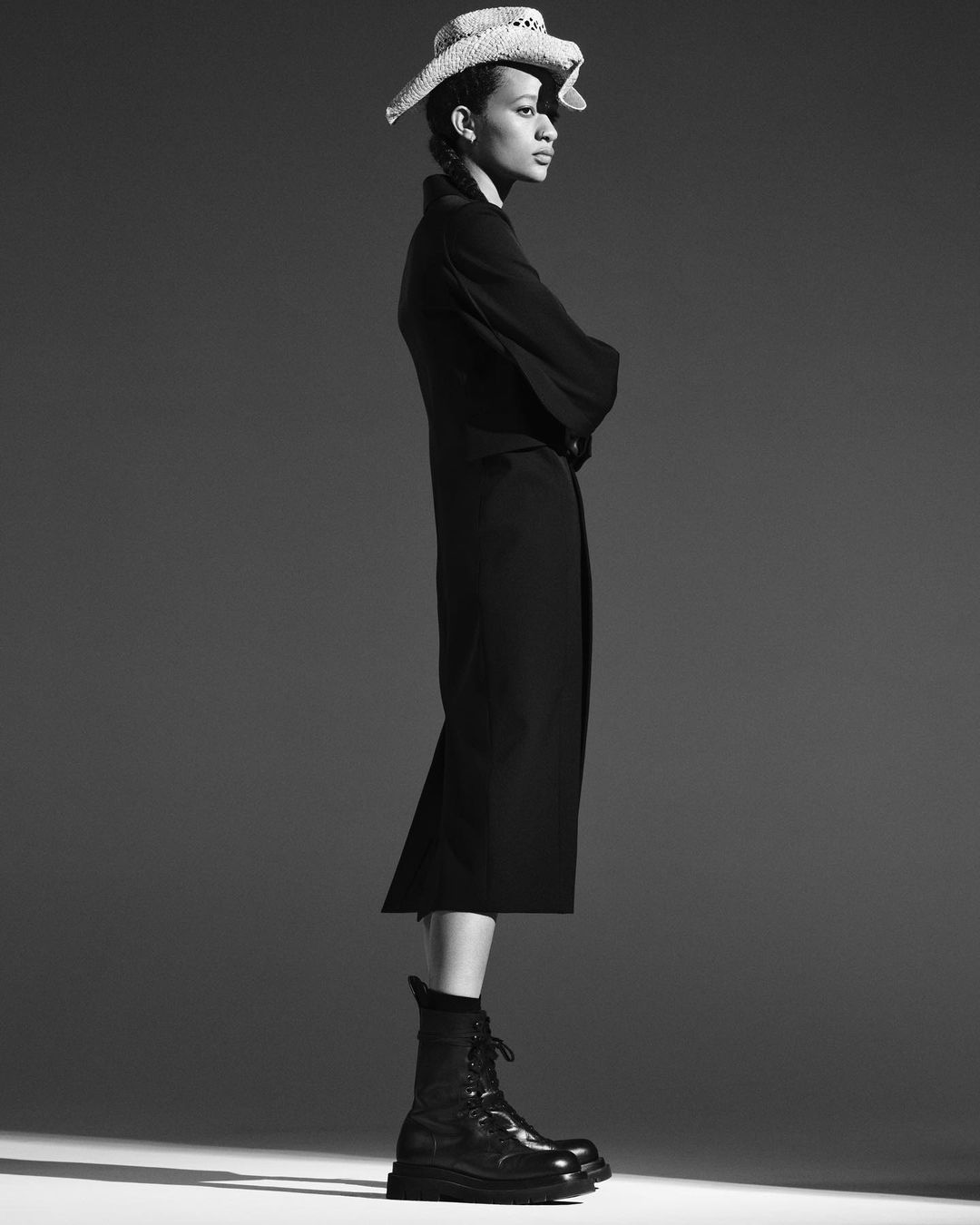 Black and white portrait of a woman wearing a long coat, boots, and straw hat, striking a confident pose in a studio setting.
