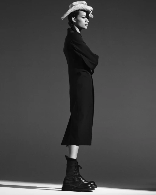 Black and white portrait of a woman wearing a long coat, boots, and straw hat, striking a confident pose in a studio setting.