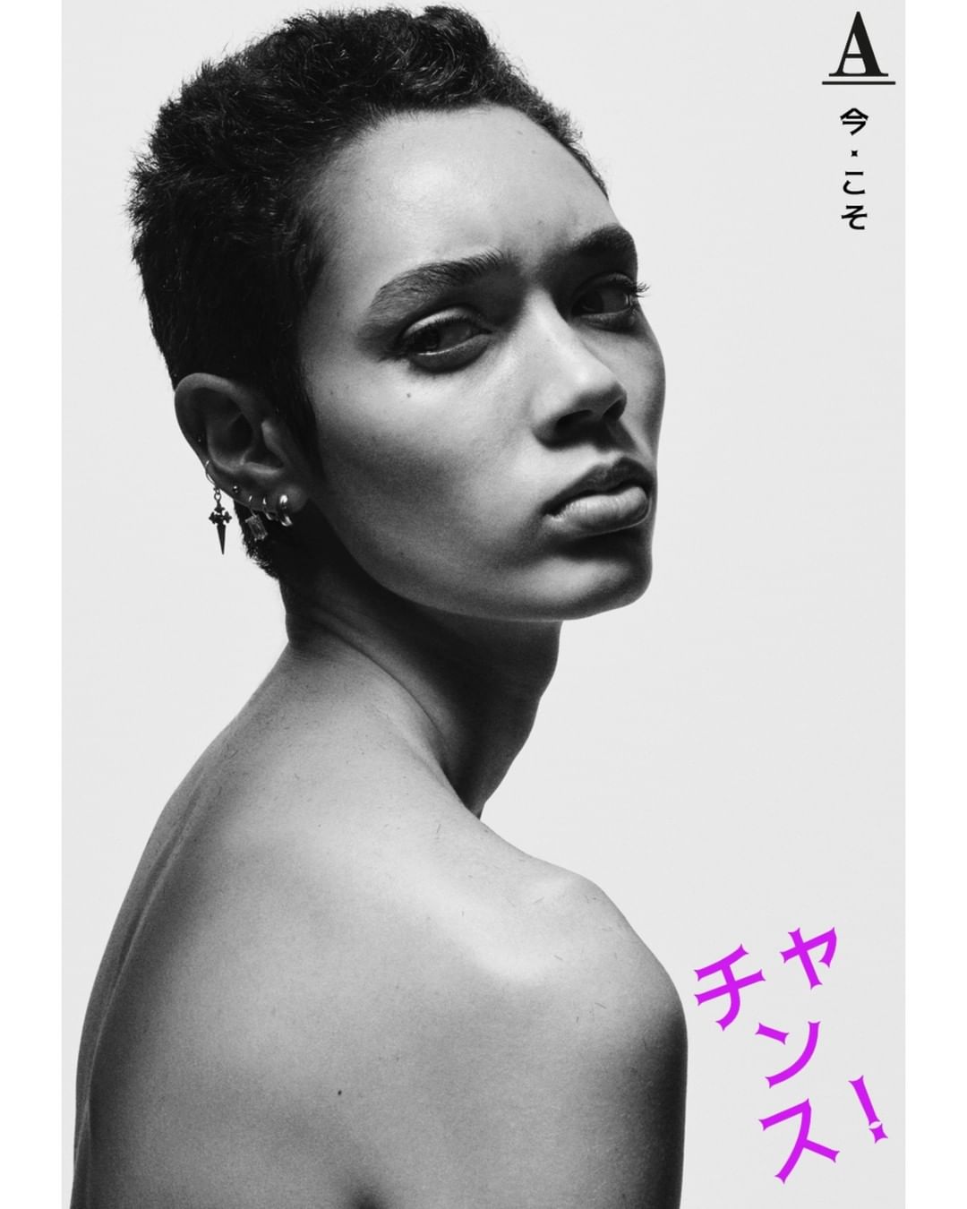 Black and white portrait of an androgynous person with short hair, wearing earrings and standing against a clean, bright white background.