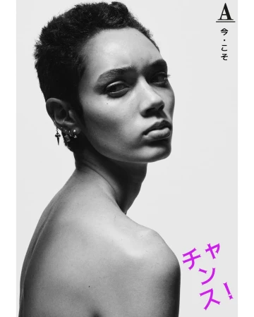 Black and white portrait of an androgynous person with short hair, wearing earrings and standing against a clean, bright white background.