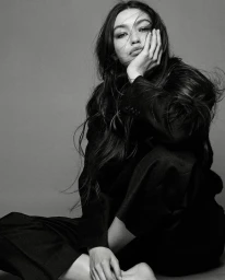 Monochrome portrait of a woman with long dark hair wearing a tailored suit, resting her cheek in her hand against a plain background.