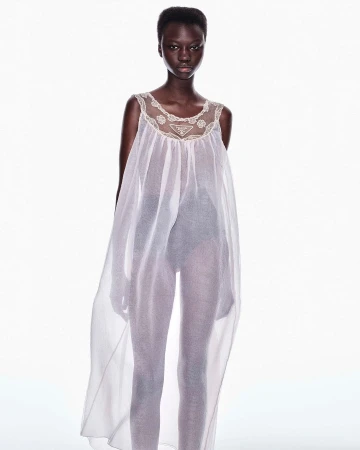 Black model standing in a sheer white sleeveless dress with lace neckline against a plain white background