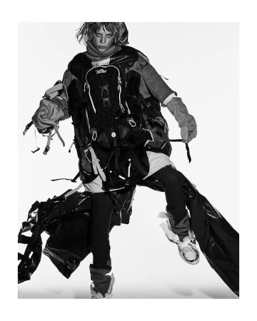 Model in layered utilitarian outerwear with backpacks and straps against a bright white seamless background